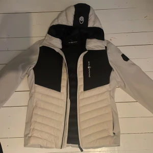 Vit och svart jacka från Sail Racing - Säljer en snygg vit och svart jacka från Sail Racing i storlek medium. Jackan är vind- och vattentät med tvåvägsstretch och dunfyllning, perfekt för kalla äventyr. Den har en cool design med logga på bröstet och en justerbar huva. Perfekt för vinterns alla äventyr! ❄️Priset är ej hugget i sten och kan sänkes till viss del. Använd cirka 2 eller 3 gånger totalt på grund av att den var för liten.