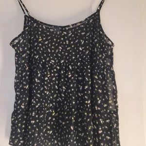 Blommig linne från Divided - Säljer ett sött blommigt linne från Divided i storlek M. Det är svart med små vita och gula blommor, perfekt för sommardagar. Linnet har smala axelband och en luftig passform. Superfint att matcha med jeans eller shorts!
