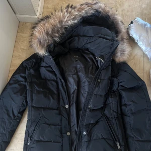 Svart dunjacka från Saki - Säljer en snygg svart dunjacka från Saki i storlek 36. Jackan har en mysig pälskantad huva och stängs med dragkedja och knappar. Perfekt för kalla vinterdagar! 🖤