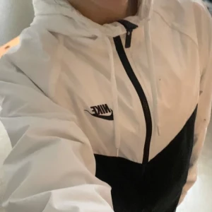 Vit och svart vindjacka från Nike - Säljer en snygg vit och svart vindjacka från Nike i storlek S. Jackan har en dragkedja framtill och är perfekt för vår och höst. Den är i mycket bra skick och har en klassisk Nike-logga på bröstet. Perfekt för både träning och vardag!
