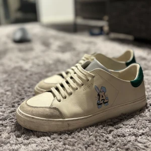 Beige sneakers, axel arigato  - Säljer ett par snygga beige sneakers från Axel Arigato med ett coolt på sidan. Skorna har en grön detalj vid hälen, bara andvända några få gånger 