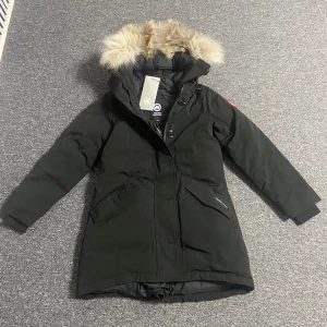 Svart victoriaparka jacka från Canada Goose - Säljer en svart parkajacka från Canada Goose i mycket bra skick. Jackan har en varm pälsfodrad huva och är perfekt för kalla vinterdagar. Den har långa ärmar och stängs med dragkedja och knappar. Märkets logga finns på ärmen. Perfekt för vintern! ❄️