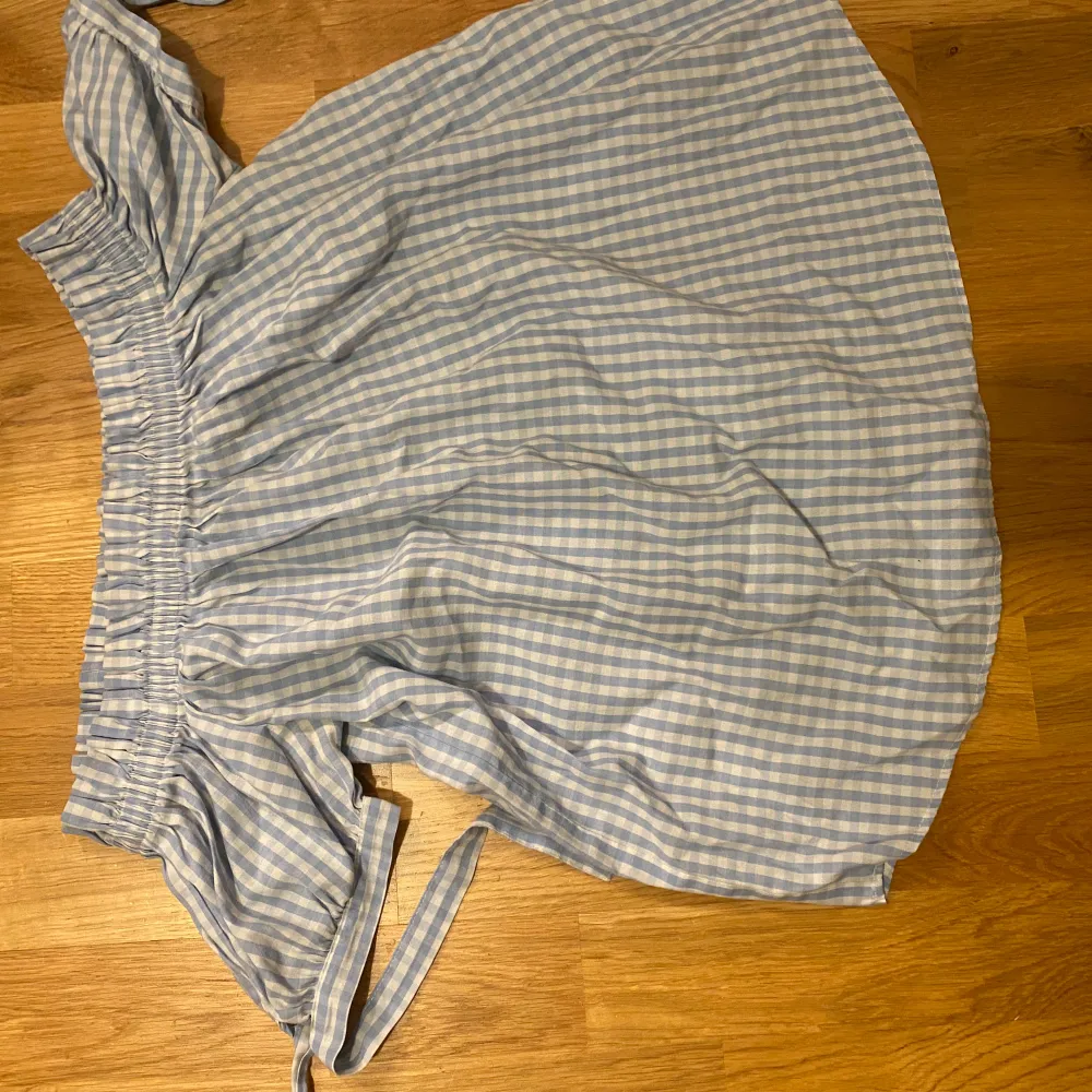 Söt blåvit rutig klänning med offshoulder-design och volangdetaljer. Perfekt för sommarens alla tillfällen! Den har en bekväm passform med elastisk midja och knytband för extra stil. Klänningen är i mycket bra skick och redo för nya äventyr.. Mekot.