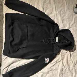 Svart hoodie från Moncler - Säljer en svart Moncler hoodie i storlek L. Den har en stor logga på ryggen i rött och blått och en mindre logga på ärmen. Perfekt för höst och vinter med sin mysiga känsla. Huvan har dragsko och det finns en stor ficka framtill. Passar både till vardags och chill.