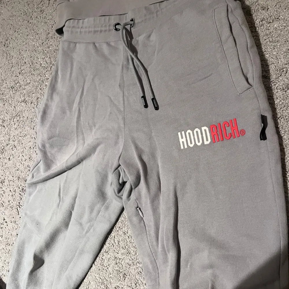 Säljer en snygg grå hoodie och matchande byxor från Hoodrich. Hoodien har en stor ficka fram och texten 'HOODRICH' i vitt och rött på bröstet. Byxorna har fickor i midjan och samma text på benet. Perfekt för en avslappnad stil! Har av er vid minsta fundering 🤔| lämna gärna prisförslag✍️. Tröjor & Koftor.