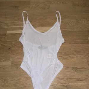 Vit ärmlös bodysuit - Säljer en stilren vit bodysuit med smala axelband och djup urringning. Perfekt för sommardagar eller som basplagg under en kavaj. Materialet är mjukt och stretchigt för en bekväm passform. Passar till både vardag och fest! genomskinlig  så man kan se vårta