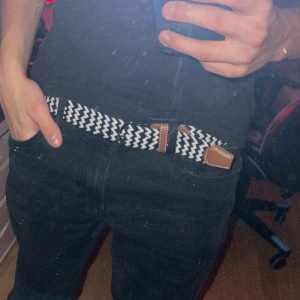 Svartvitt flätat bälte - Säljer ett snyggt svartvitt flätat bälte med bruna läderdetaljer. Perfekt för att ge lite extra stil till din outfit. Bältet har en klassisk design med ett mönster som verkligen sticker ut. Passar både till vardags och fest!