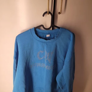 Blå sweatshirt med strassdetaljer - Säljer en snygg blå sweatshirt i storlek M. Tröjan har coola strassdetaljer på framsidan som ger den en unik look. Perfekt för en avslappnad stil eller att glamma upp vardagen. Den är i bra skick och redo för nya äventyr!