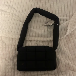 Svart quiltad axelväska - Snygg svart quiltad axelväska i syntetmaterial. Perfekt för både vardag och fest med sitt stilrena och moderna utseende. Väskan har en bred axelrem som gör den bekväm att bära. Enkelt att matcha med olika outfits!