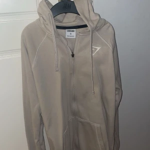 Beige zip hoodie från Gymshark - Säljer en beige hoodie från Gymshark i storlek xs. Den har dragkedja framtill och en praktisk huva med dragsko. Perfekt för både träning och vardag. Hoodien är i mycket bra skick och superbekväm att ha på sig. Tröjan är dessutom aldrig använd!