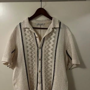 Vit kortärmad stickad / virkad piké från Zara - Säljer en snygg vit skjorta från Zara i storlek M. Den har ett coolt stickat mönster med svarta detaljer och knappar framtill. Perfekt för sommardagar eller en avslappnad fest. Skjortan är i mycket bra skick och ger en stilren look.