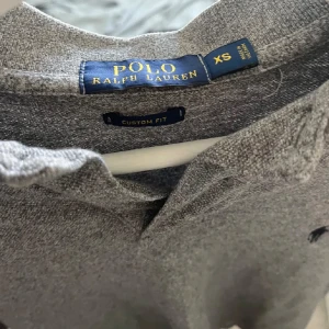 Grå pikétröja från Ralph Lauren - Säljer en stilren grå pikétröja från Ralph Lauren i storlek XS. Tröjan har en klassisk krage med två knappar och det ikoniska logotypbroderiet på bröstet. Perfekt för både vardag och lite finare tillfällen. Passar bra till jeans eller chinos!
