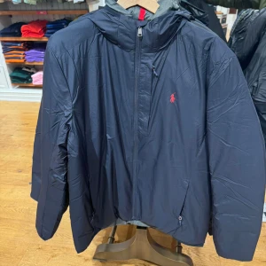 Svart packbar jacka från Ralph Lauren - Säljer en svart, lätt och packbar jacka från Ralph Lauren. Perfekt för höst och vår med en praktisk dragkedja och huva. Jackan har en liten röd logga på bröstet och kan enkelt packas ihop för smidig förvaring. Perfekt för resor eller som en extra jacka i väskan!