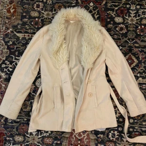 Beige mockakappa med fuskpäls! - Säljer en snygg beige mockajacka med fuskpäls på kragen. Jackan har långa ärmar och knäpps med knappar framtill. Den har också ett bälte i midjan för en mer figurnära passform. Perfekt för höst och vinter! Den har mysigt tyg på insidan som är skönt 🤍 Längst ner på kappan har insidans tyg lossnat lite, syns på sista bilden, men det går enkelt att fixa med nål och tråd!