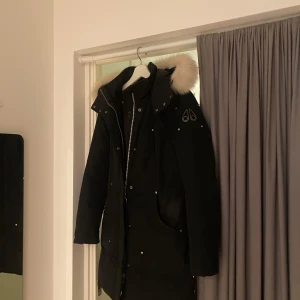 Svart parkajacka från Moose Knuckles - Säljer en snygg svart lång parkajacka från Moose Knuckles i storlek S. Jackan har en fluffig vit pälskant på huvan och är perfekt för kalla vinterdagar. Den har dragkedja och knappar framtill för extra skydd mot kylan.  Köpt helt ny på johnells för 11 500kr och sparsamt använd 3 vinter säsonger. Inga slitatge. Tyvärr medföljer inget kvitto av jackan eftersom att jag inte kan hitta de. För frågor och fler bilder är det bara att höra av sig. 