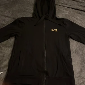 Svart hoodie från Emporio Armani - Säljer en stilren svart hoodie från Emporio Armani EA7. Den har en dragkedja framtill och en bekväm huva. Perfekt för både vardag och träning. Passar bra till höst och vinter.
