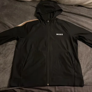 Svart huvtröja från BOSS - Säljer en snygg svart huvtröja från BOSS med dragkedja och huva. Den har en vit och guldig rand på ärmen och BOSS-loggan på bröstet. Perfekt för en casual look eller träning. Tröjan är i mycket bra skick och redo för nya äventyr! Nypris 1700kr