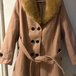 Beige kappa med fuskpäls - Säljer en superfin beige kappa med fuskpäls på kragen. Den har stora svarta knappar och ett bälte i midjan. Perfekt för höst och vinter! 🧥✨