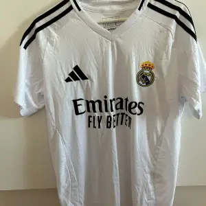 Säljer en vit Real Madrid fotbollströja från Adidas i nytt skick. Tröjan har korta ärmar och är prydd med klubbens emblem och sponsorlogga 'Emirates Fly Better'. Den är tillverkad i ett lätt och andningsbart material, perfekt för både träning och match.