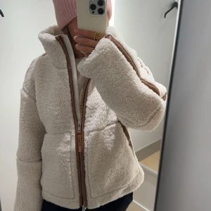 Vit teddyjacka med dragkedja - Supermysig vit teddyjacka med dragkedja och bruna detaljer. Perfekt för kyliga dagar när du vill vara både varm och stilren. Jackan har en hög krage och långa ärmar, vilket gör den idealisk för höst och vinter. Den är i mycket bra skick och storleken är XS💕