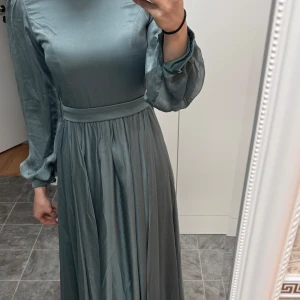 Grå långklänning med långa ärmar - Elegant grå långklänning i ett mjukt och glansigt material. Klänningen har långa ärmar med en lätt puffig stil och en markerad midja som ger en fin siluett. Är 166cm och klänningen är i storlek 36