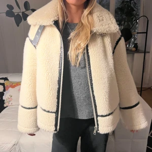 Vit teddyjacka med svarta detaljer - Supermysig vit teddyjacka med svarta detaljer och dragkedja framtill. Perfekt för kyliga dagar med sin fluffiga och varma design. Använd ungefär 3 gånger men köptes förra året. Helt nyskick. Nypris 1200