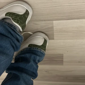 Vita sneakers med gröna och röda detaljer - Säljer ett par vita sneakers med gröna och röda detaljer. Skorna har en cool krokodiltextur på vissa delar och klassisk snörning. De är i bra skick men har lite slitage på sulorna. Perfekta för en avslappnad stil!