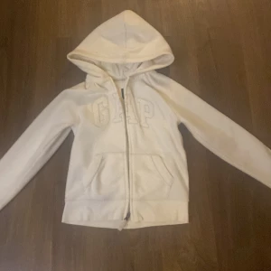 Vit hoodie från GapKids - Säljer en superfin vit hoodie från GapKids i storlek 10-11 år. Den har en dragkedja framtill och en mysig huva. GAP-loggan på bröstet är glittrig och ger en extra touch. Perfekt för höst och vår! Den är väldigt mysig och nästan lite foder inuti.