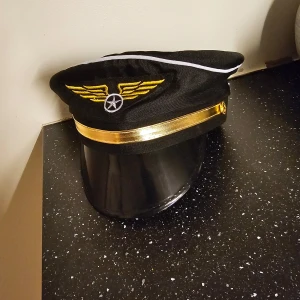 Svart pilotmössa med gulddetaljer - Snygg svart pilotmössa med guldfärgad skärm och broderade vingar framtill. Perfekt för temafester eller som en cool accessoar. Mössan har en vit kantlinje som ger en extra touch av stil.