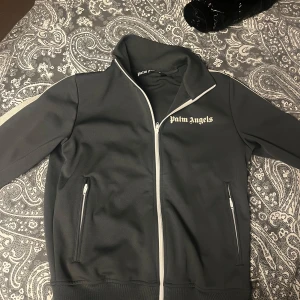 Grå track jacket från Palm Angels - Säljer en snygg grå track jacket från Palm Angels. Den har en vit dragkedja och vita ränder längs ärmarna. Jackan är i mycket bra skick och perfekt för både vardag och fest. Den har två fickor med dragkedjor framtill. Kom gärna med bud