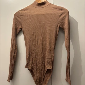 Beige ribbad bodysuit - Säljer en snygg beige ribbad bodysuit i storlek XS. Den har långa ärmar och en hög krage, perfekt för lager-på-lager under höst och vinter. Materialet är mjukt och stretchigt, vilket gör den bekväm att bära hela dagen🤎