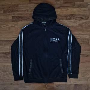 Säljer en snygg oanvänd hoodie från Hugo Boss i mycket bra skick. Den har en dragkedja framtill och en huva för extra komfort. Perfekt för höst och vår, med vita detaljer längs ärmarna som ger en sportig look. Det är en eprfekt julklapp åt dig själv eller nära och kära🎁 Passa på!