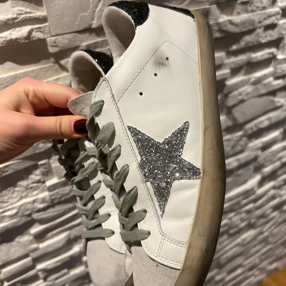 Säljer ett par snygga Golden Goose sneakers i vitt skinn med en glittrig silverstjärna på sidan. Skorna har grå snörning och svart glittrig häl. Perfekta för att ge din outfit lite extra bling! . Skor.