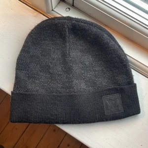 Svart mössa med logotyp - Snygg svart mössa med en diskret logotyp framtill. Perfekt för kalla dagar och ger en stilren look. Materialet ser ut att vara mjukt och bekvämt, vilket gör den idealisk för både vardag och speciella tillfällen.