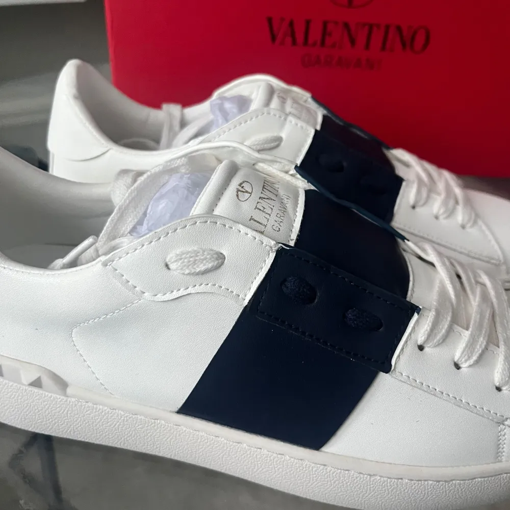 Säljer ett par snygga Valentino Garavani sneakers i vitt skinn med en bred blå rem över snörningen. Skorna har den klassiska rockstud-detaljen på hälen och en mönstrad sula för extra grepp. Kommer i originalkartong. Storlek 43 ☺️. Skor.