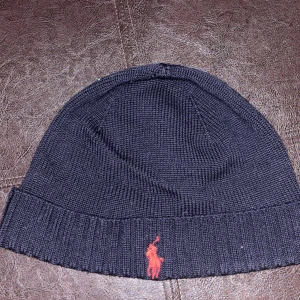 Mörkblå stickad mössa från Polo Ralph Lauren - Snygg mörkblå stickad mössa från Polo Ralph Lauren med röd broderad logga framtill. Perfekt för att hålla värmen under kalla dagar. Den har en ribbad kant som ger en skön passform. Passar till både vardag och mer uppklädda tillfällen.