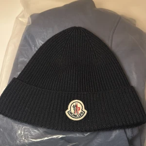 Svart  mössa från Moncler i färgen marinblå - Säljer en snygg svart mössa från Moncler. Den är i nytt skick och har en broderad logga framtill. Perfekt för kalla dagar och kommer se många mössor moncler dena vintern. Passar till både vardag och fest!