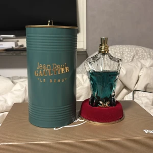 Jean Paul Gaultier Le Beau Parfym 125ml - Säljer en elegant parfymflaska från Jean Paul Gaultier, 'Le Beau'. Flaskan har en unik design som liknar en manlig torso och innehåller en blågrön vätska. Den kommer i en stilren grön metallförpackning med guldtext. Perfekt för den som vill ha en lyxig och iögonfallande doft. Pris kan diskuteras