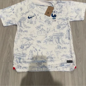 Vit och blå fotbollströja från Nike - Snygg vit fotbollströja från Nike med blå detaljer och ett unikt mönster. Tröjan har korta ärmar och en stilren design med en blå Nike-logga och emblem på bröstet. Perfekt för fotbollsfans som vill sticka ut på planen!