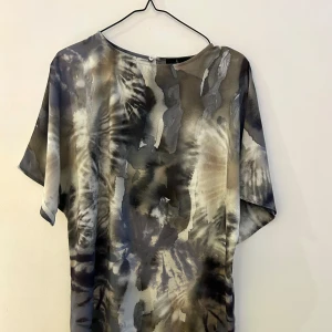 Silkig klänning / top - Snygg klänning från Åhléns med abstrakt mönster i grå och beige vattenfärgsliknande toner. Den har en lös passform och korta ärmar och passar lika bra som klänning som till top över smala byxor eller jeans. Materialet i 100% återvunnen polyester är mjukt och bekvämt.