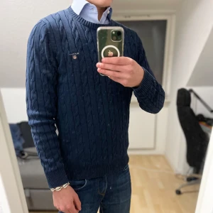 Kabelstickad tröja från GANT - Säljer en stilren mörkblå kabelstickad tröja från GANT. Tröjan har en klassisk rund halsringning och ett diskret broderat emblem på bröstet. Perfekt för en elegant och avslappnad look. Jätte bra nu för dem kallare tiderna där man vill klä upp sig lite🤵🏼‍♂️