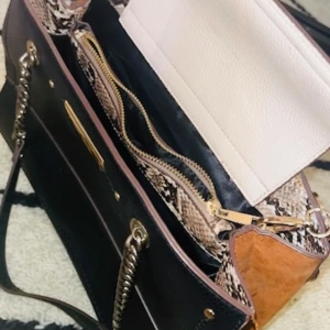 Svart och brun axelväska med kedjedetaljer - Snygg svart och brun axelväska från River Island med kedjedetaljer och ett stilrent mönster på insidan. Väskan har flera fack och en dragkedja för säker förvaring. Perfekt för att bära med sig det viktigaste med stil.