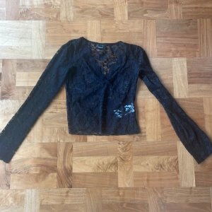 Svart spetstopp från Gina Tricot  - Snygg svart spetstopp från Gina Tricot med v-ringning och långa ärmar. Perfekt för en elegant look. Toppen har ett vackert spetsmönster som ger en romantisk touch.