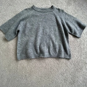 Grå stickad tröja från 157 - Mysig grå stickad tröja från 157 i storlek XS. Tröjan har korta ärmar och en rund halsringning. Perfekt för kyligare dagar.