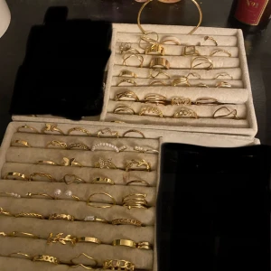 Samling av guldiga ringar och armband - En elegant samling av guldiga ringar och armband. Ringarna har olika design, inklusive enkla band, mönstrade och med små dekorationer som pärlor och stenar. Armbanden är i matchande guldtoner, perfekta för att komplettera din stil. Får all på bilden.