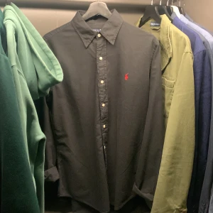 Svart skjorta från Ralph Lauren - Snygg svart skjorta från Ralph Lauren med klassisk krage och knappar framtill. Den har en röd broderad logotyp på bröstet och är perfekt både till sommaren och vardagen. Den kommer tyvärr aldrig till användning och säljer därför. Nypris ligger runt 1500 men mitt pris ligger runt 700 