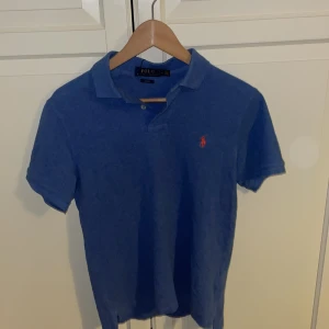 Blå pikétröja från Ralph Lauren - Säljer en klassisk blå pikétröja från Ralph Lauren med korta ärmar och den ikoniska loggan broderad i rött på bröstet. Perfekt för en stilren och avslappnad look. Inga defekter och ett väldigt bra skick pris kan diskuteras 