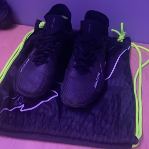 Svarta fotbollsskor från Nike - Snygga svarta Nike fotbollsskor med snörning och Air Zoom-teknologi. Skorna har en modern design med diskreta detaljer och kommer med en praktisk svart och neongrön påse.