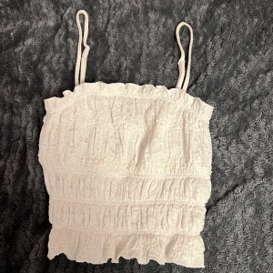 Vit axelbandslös topp - Söt vit axelbandslös topp med smala axelband och volangdetaljer. Perfekt för sommardagar och kan enkelt matchas med olika outfits.