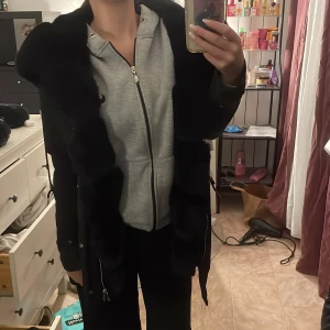 Svart pälsjacka med dragkedja - Snygg svart pälsjacka med dragkedja och långa ärmar. Jackan har en lyxig känsla med mjuk pälsdetalj längs framsidan. Perfekt för att hålla sig varm och stilren under kyliga dagar.